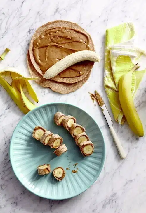 Preparación de banana sushi con tortilla integral, mantequilla de maní y rodajas de plátano en un plato azul.