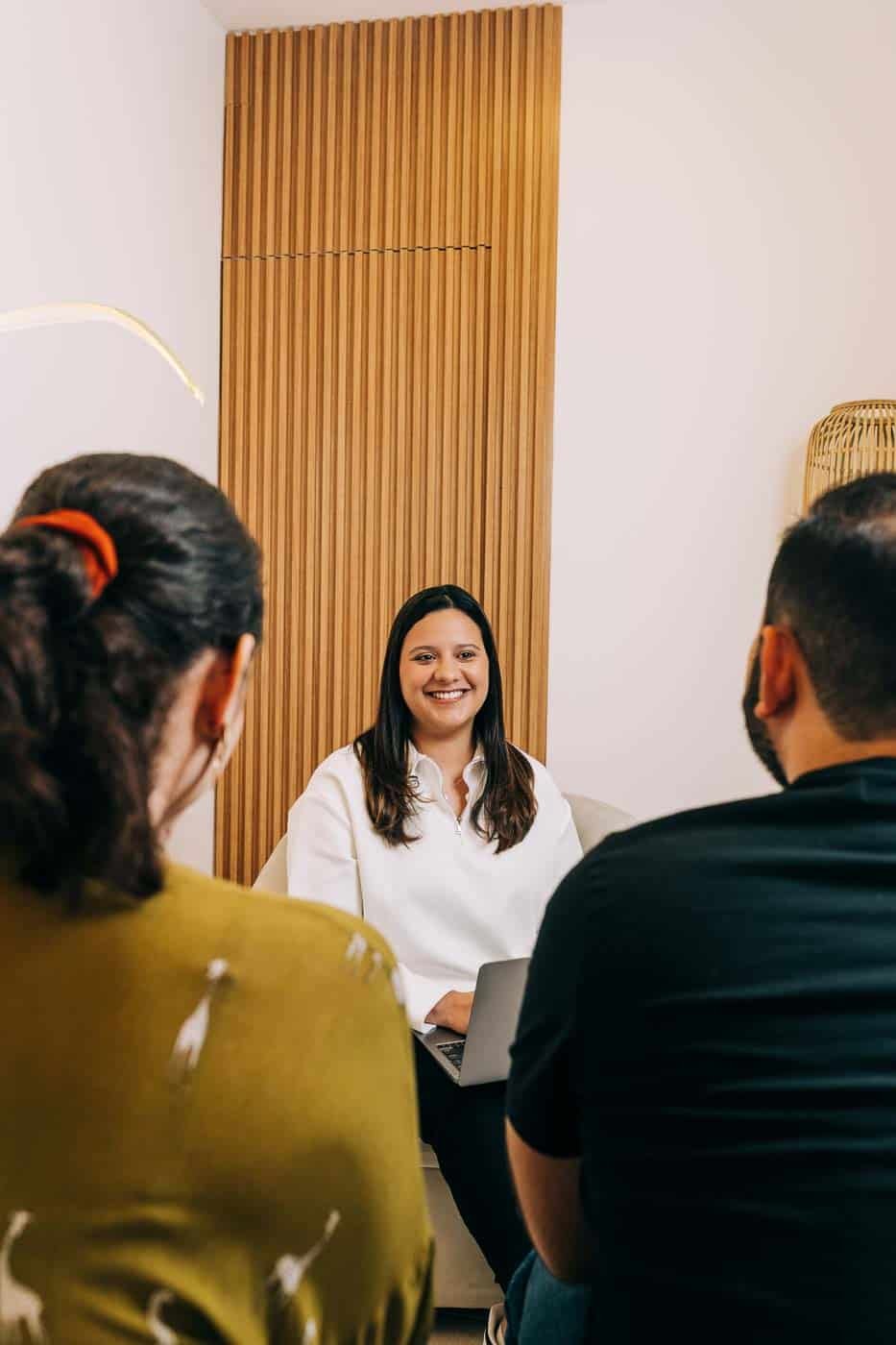 Terapia de pareja en sesión con la psicóloga Rebeca Martínez en El Salvador. Rebeca sonríe mientras conversa con una pareja sentada frente a ella en un ambiente cálido y profesional.