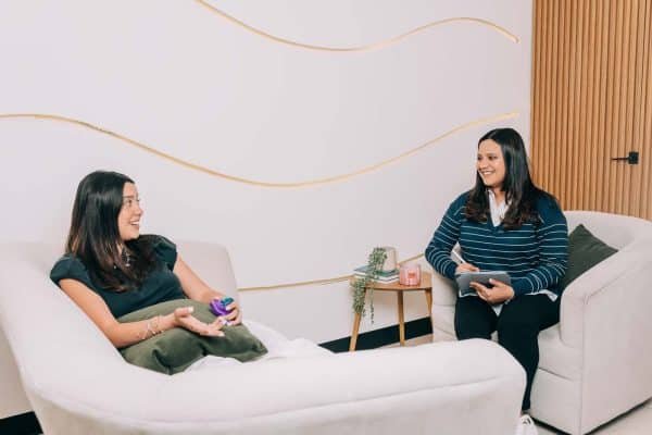 Psicóloga Rebeca Martínez conversando con una paciente en una sesión de terapia en un entorno cómodo y acogedor.