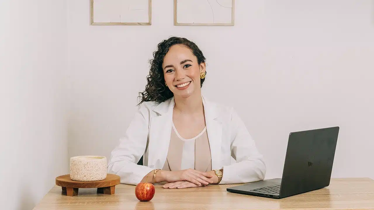 Gabriela Baranoa, nutricionista en Mood Clinic, sonríe sentada en un escritorio con una computadora portátil negra, una manzana roja y una vela decorativa en un ambiente minimalista y profesional.