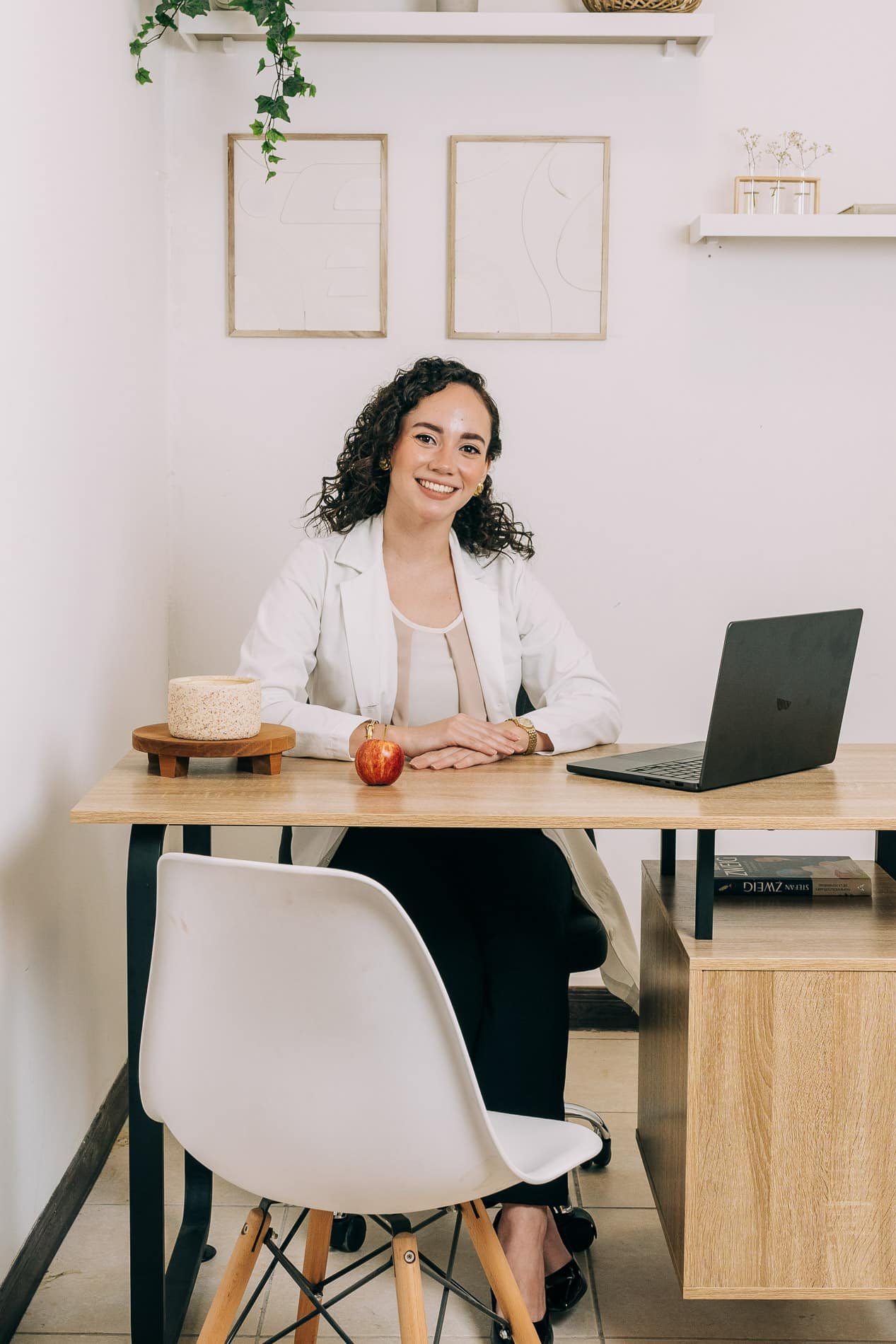 Nutricionista Gabriela sentada en su oficina con una manzana roja sobre su escritorio, rodeada de decoración minimalista y una computadora portátil.