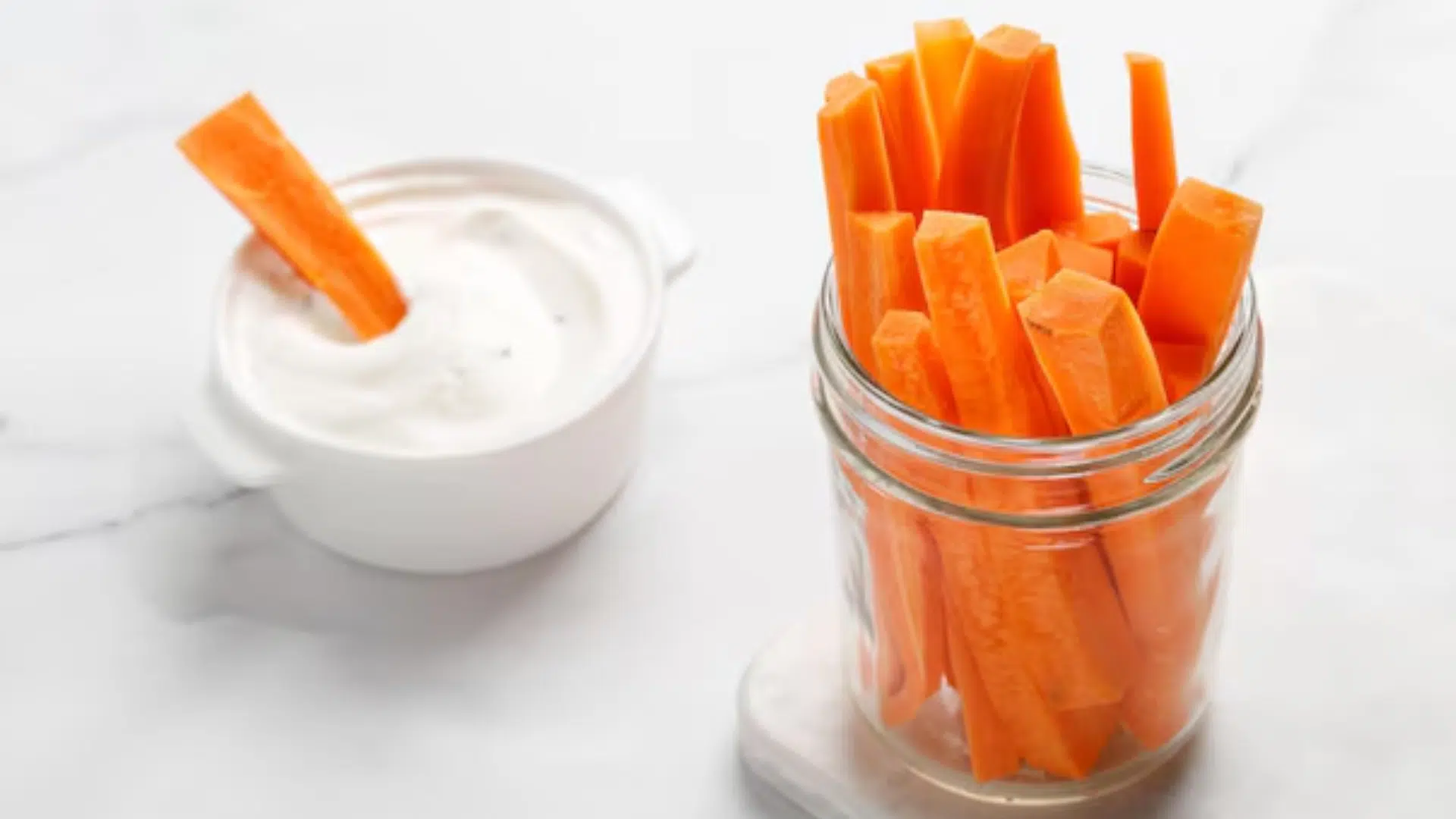 Snack saludable de bastones de zanahoria servidos en un frasco de vidrio junto a un dip blanco en un recipiente pequeño.