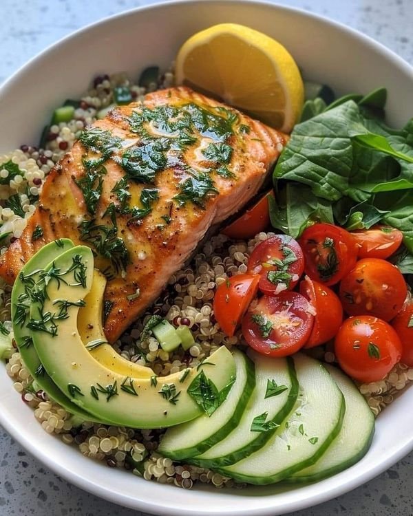 Plato de salmón a la parrilla con quinoa, aguacate, espinacas, tomate cherry y pepino, decorado con hierbas frescas y rodaja de limón.